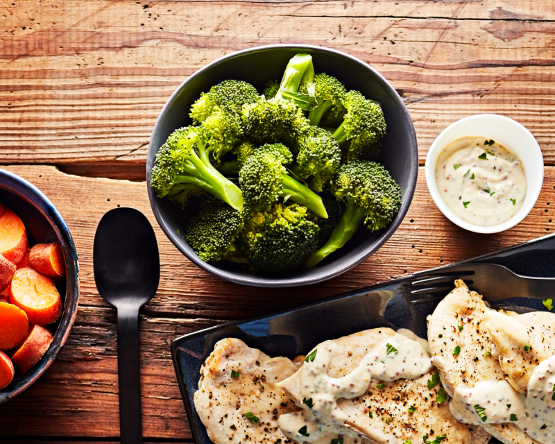 A close up of a bowl of steamed broccoli with a plate of grilled chicken breast and bowl of steamed carrots