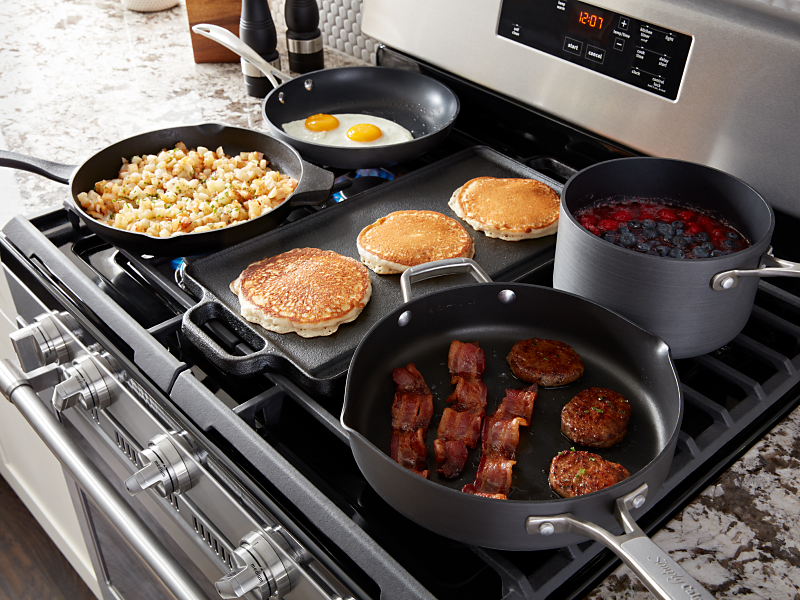 Various breakfast foods cooking on a gas stove and griddle