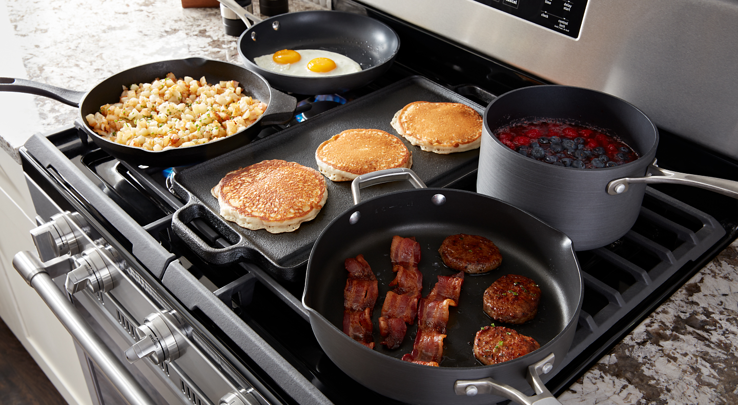 Various breakfast foods cooking on a gas stove and griddle