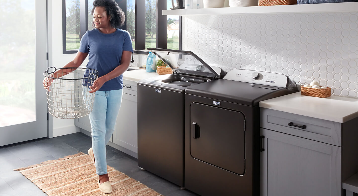 A woman holding a laundry basket next to Maytag® side-by-side washer and dryer set in a modern laundry room