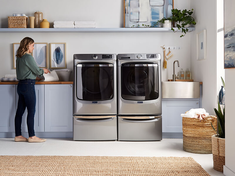 A woman folding laundry next to Maytag® side-by-side washer and dryer set in a modern laundry room