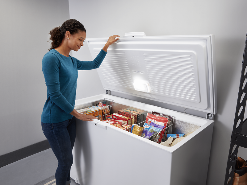 A woman opening a white chest freezer with various frozen items inside