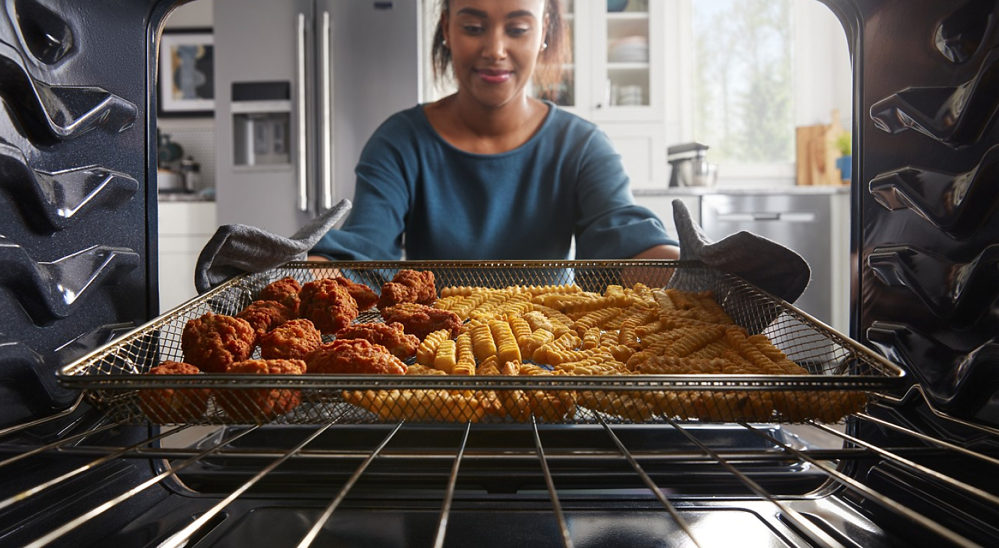 A woman pulling an air fry basket filled with fries and chicken wings out of the oven.