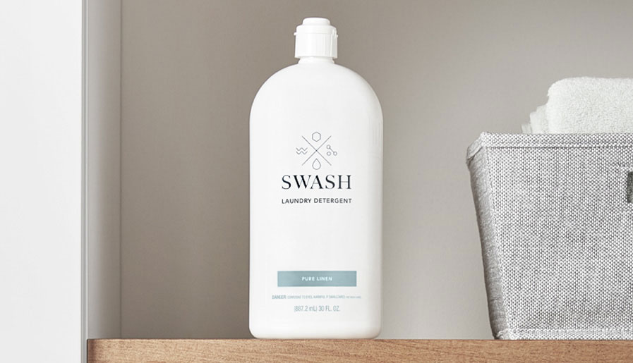 Du détergent à lessive Swash Linge frais sur une tablette en bois dans une salle de lavage, près de serviettes-éponges et autres articles de lessive