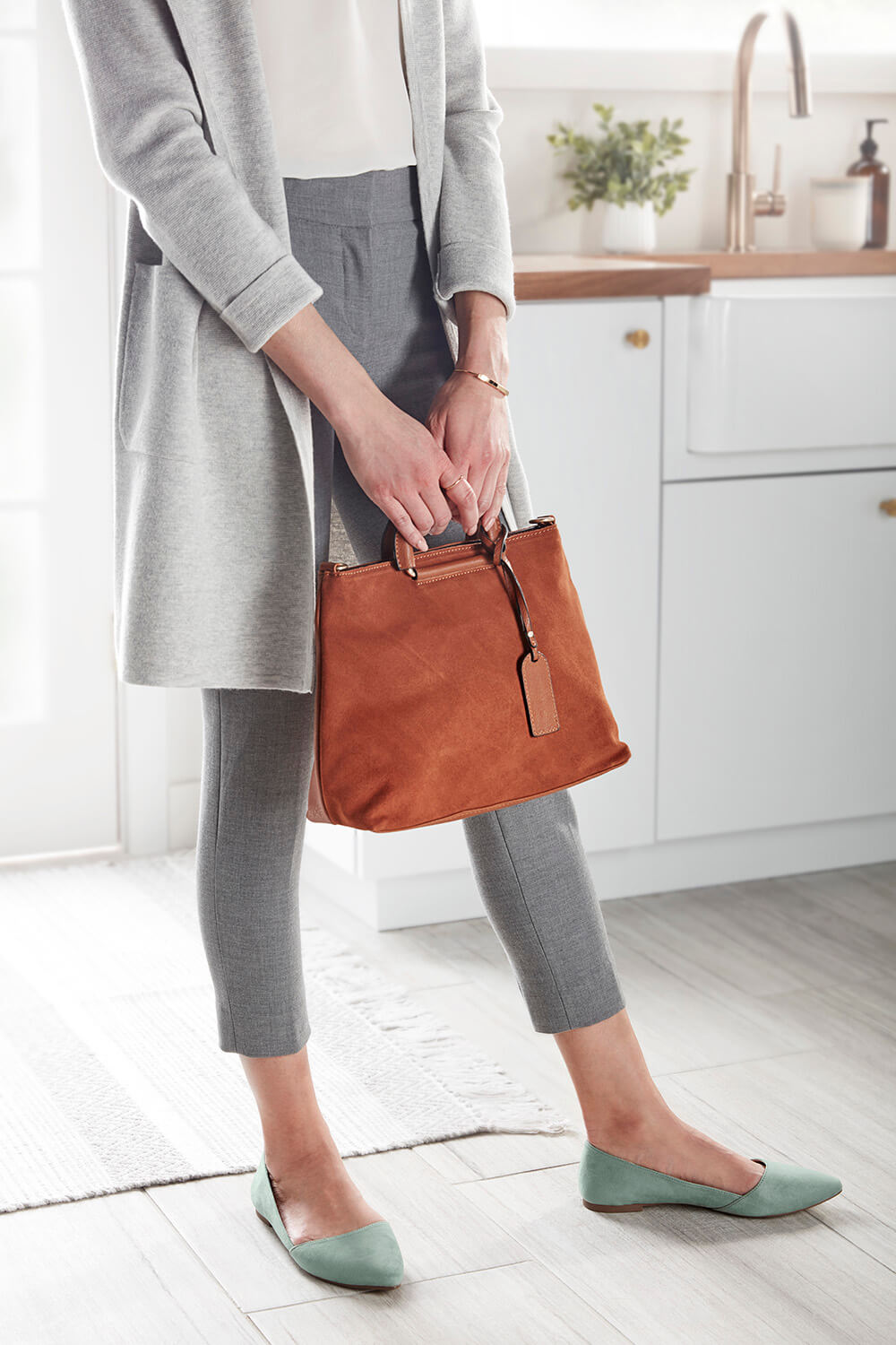 A woman dressed in gray pants, holding an orange purse in her hands, standing in her kitchen