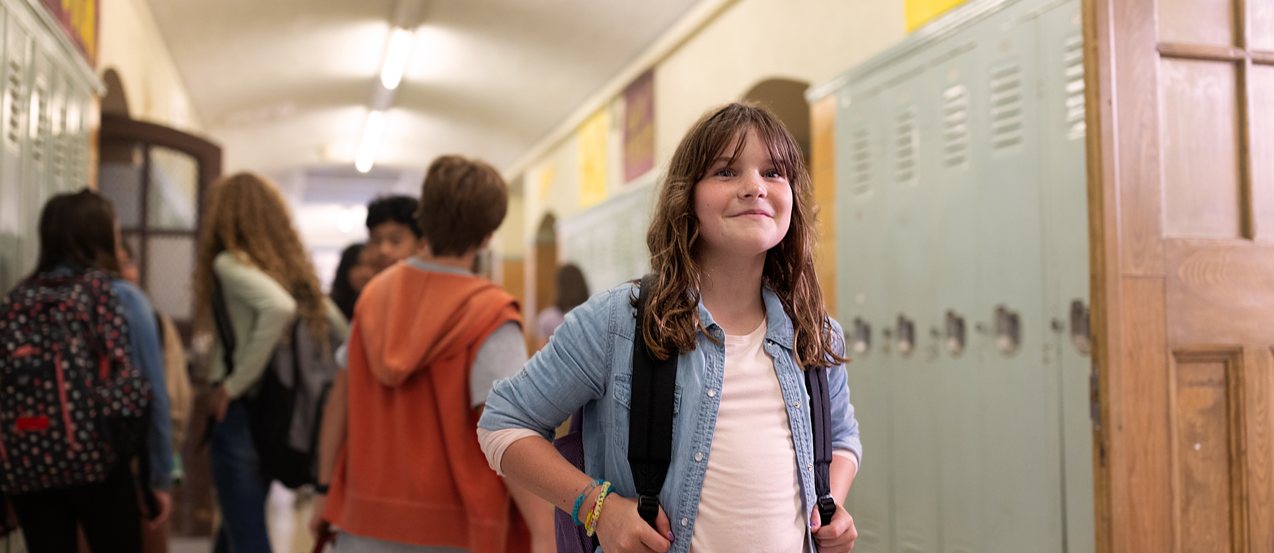 A smiling child in a school hallway with other kids behind them