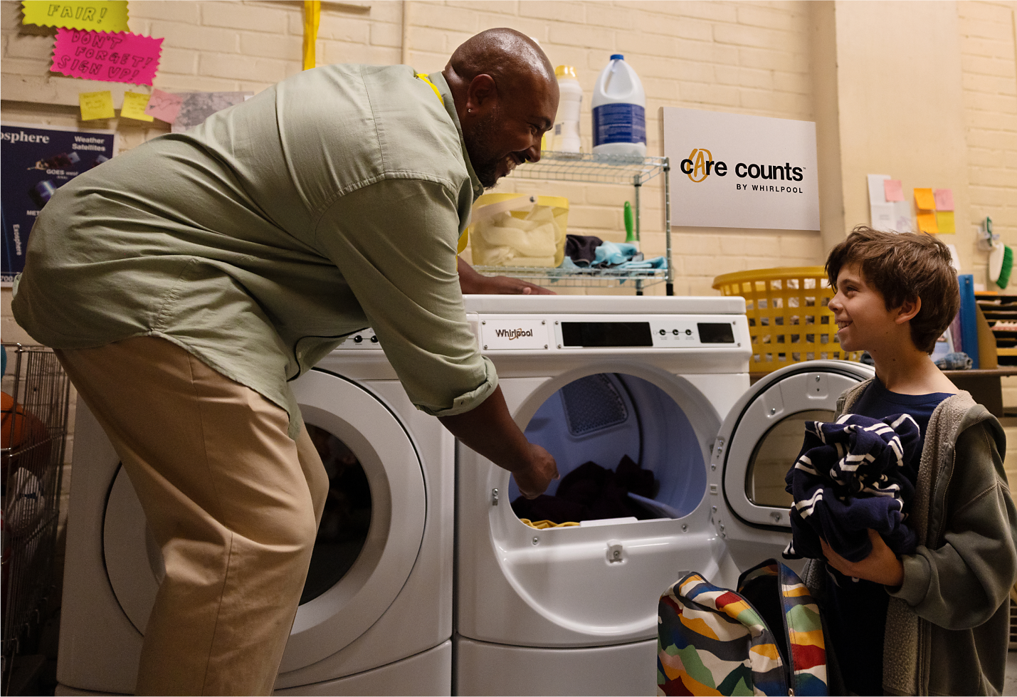 An adult and a child load clothes into a Whirlpool® Dryer in a school