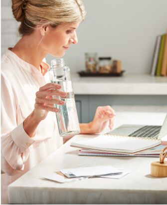 Une femme boit dans une bouteille d'eau réutilisable remplie d'eau claire tout en travaillant sur l'ordinateur