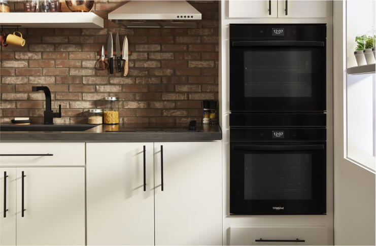 A Whirlpool® Small Space Double Wall Oven, Electric Cooktop and Wall Mount Range Hood in a kitchen with light cabinets