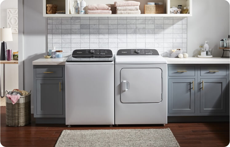 A white top load Whirlpool® Washer and Dryer in a laundry room with gray cabinets. 