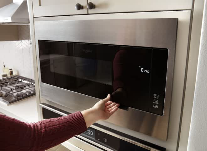 An arm reaches for the handle of a Whirlpool® Built-In Microwave