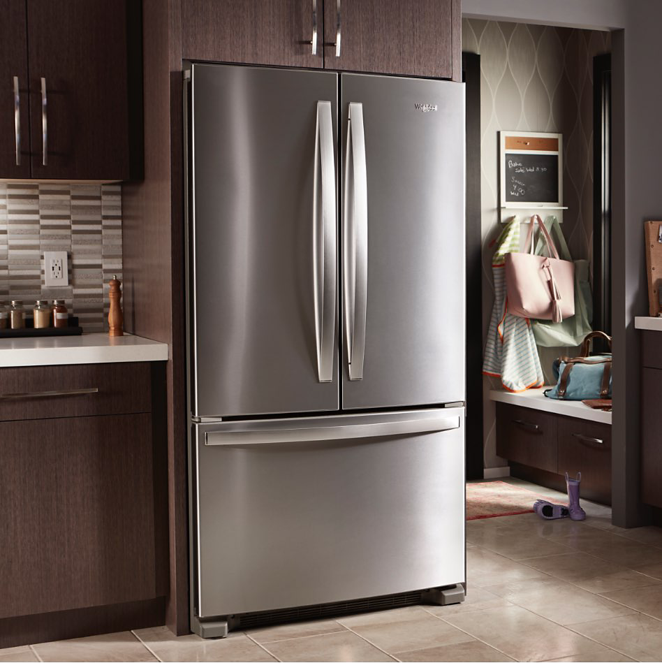 A stainless steel side-by-side refrigerator next to a brown cabinet and drawer. On the counter is a line of jarred spices in front of a grey and white backsplash with an outlet. Above are two brown cabinets