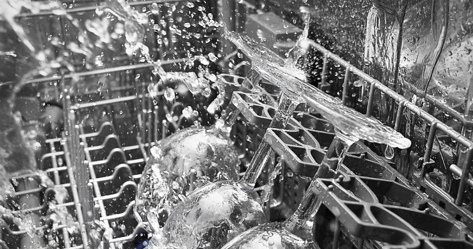 The interior of a dishwasher during a wash cycle. Water is spraying everywhere and there are three wine glasses on the rack.