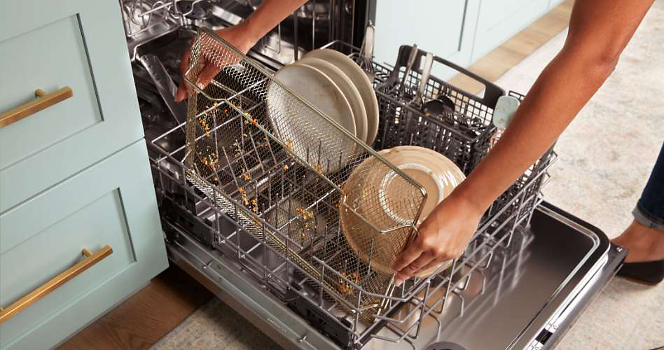 Someone loading an air fry basket into a Whirlpool dishwasher. On the lower rack are plates, a bowl and a cutlery tray.