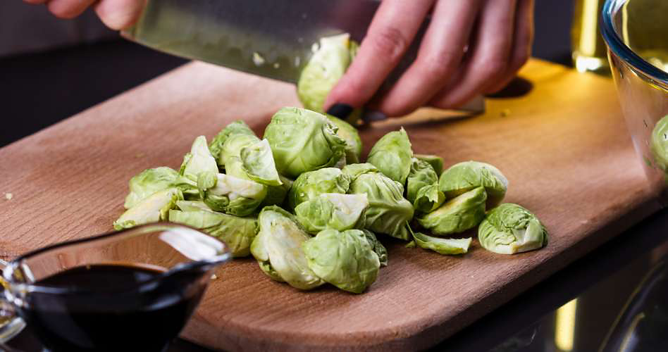 Someone slicing Brussels sprouts on a cutting board. Next to the cutting board is a small bowl of sauce.
