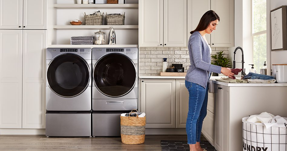Inside a laundry room. Next to white cabinets are a washer and dryer. Above the washer and dryer are shelves filled with jars and baskets. On the floor, in front of the washer and dryer, is a wicker basket. A woman in a grey cardigan and jeans folds dish cloths next to a sink. Next to her is a white hamper filled with towels and/or dish cloths.