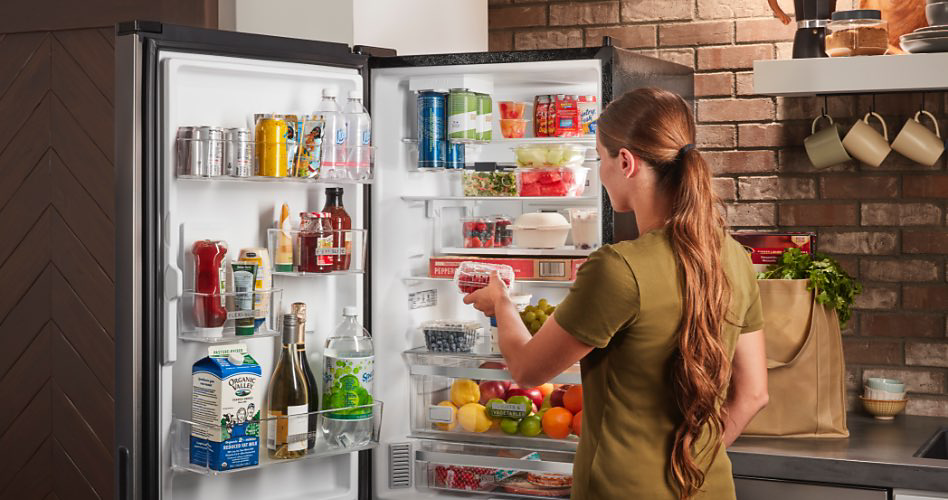 Woman stands in front of opened Whirlpool fridge holding a carton. Inside are beverage cans, water bottles, ketchup, mayo, carton of milk, wine, and various produce.