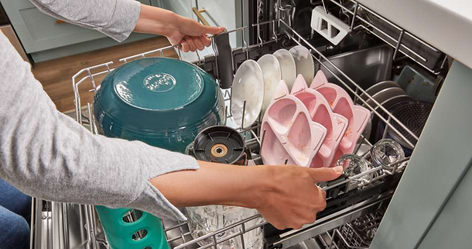 Someone adjusts the positioning of a rack in a Whirlpool dishwasher. On the rack are a large ceramic bowl, plates, pink compartmentalized plates for kids and glasses