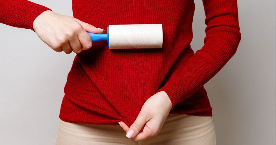 A person in a red sweater and beige pants pulls their sweater down with one hand and is about to remove lint with a lint roller