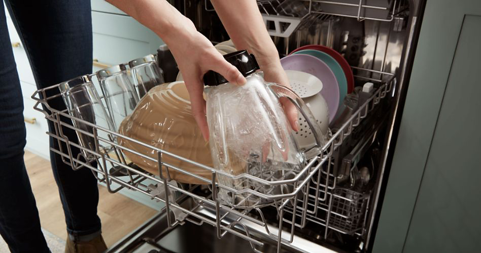 Someone touching a blender jar that's in a dishwasher rack. Also on the rack are a ceramic bowl, plates and a colander.
