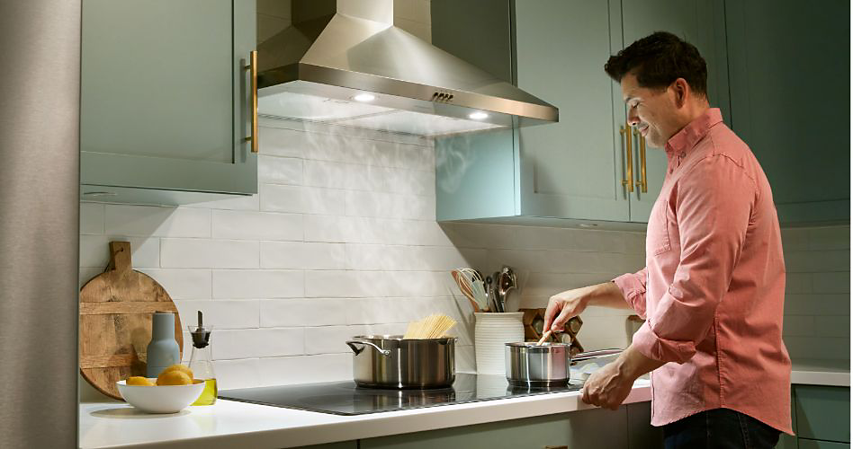 A man cooks at a Whirlpool Range with a Range Hood. There's a pot of pasta on the stove. On the counter are a bowl of fruit, a container of kitchen utensils, an oil container and a cutting board leans against the wall