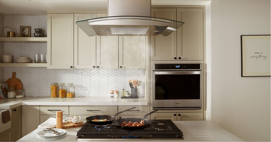 A kitchen with a Whirlpool Wall Oven and a Whirlpool Cooktop with a Range Hood. On the counters are a cutting board, containers of flour, containers of pasta, a container of kitchen utensils and shelves with plates, glasses and more