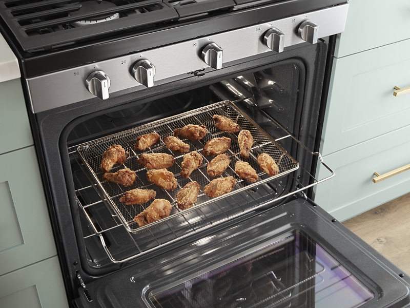An open oven door with an air fryer basket containing chicken wings slid out.