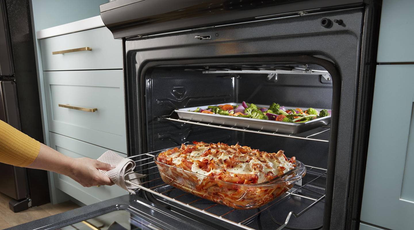 Person pulling out lasagna from the low oven rack while vegetables cook on the top rack