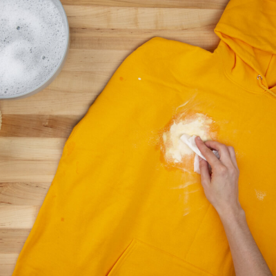 Person using a spoon to scrape off dried white paint from a yellow sweatshirt
