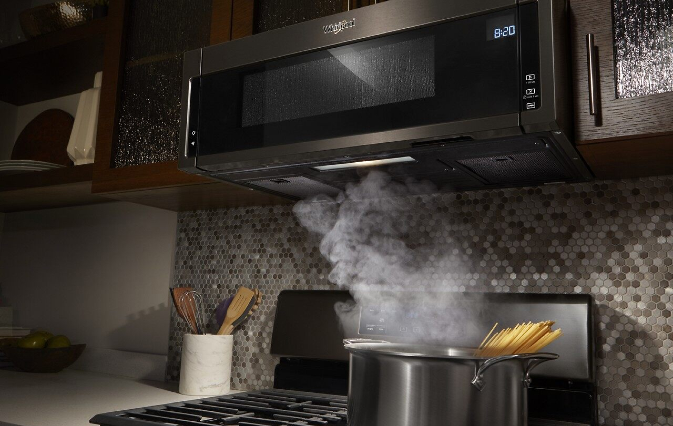 Silver and black over-the-range microwave installed with pasta steaming on the stove below