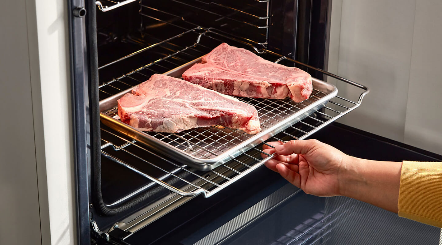 A person sliding a rack of raw steaks into an oven