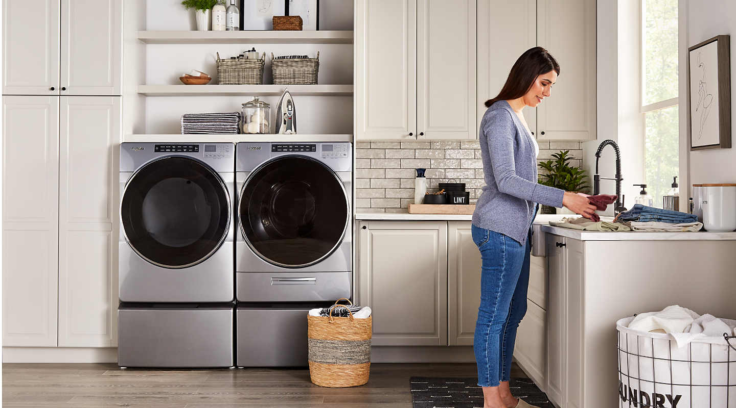 A person folding laundry with a Whirlpool® washer and dryer nearby. A person folding laundry with a Whirlpool® washer and dryer nearby.