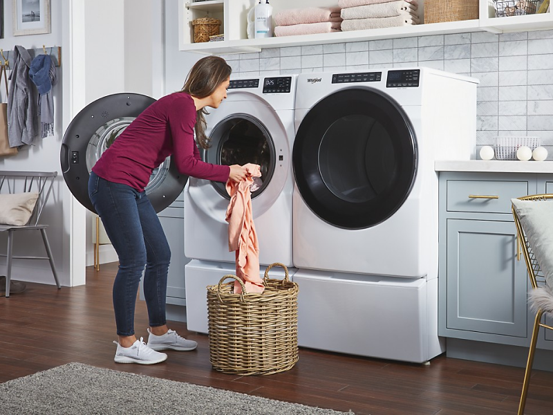 A woman putting laundry into a white Whirlpool® front load washer that sits next to a dryer in a modern laundry room.