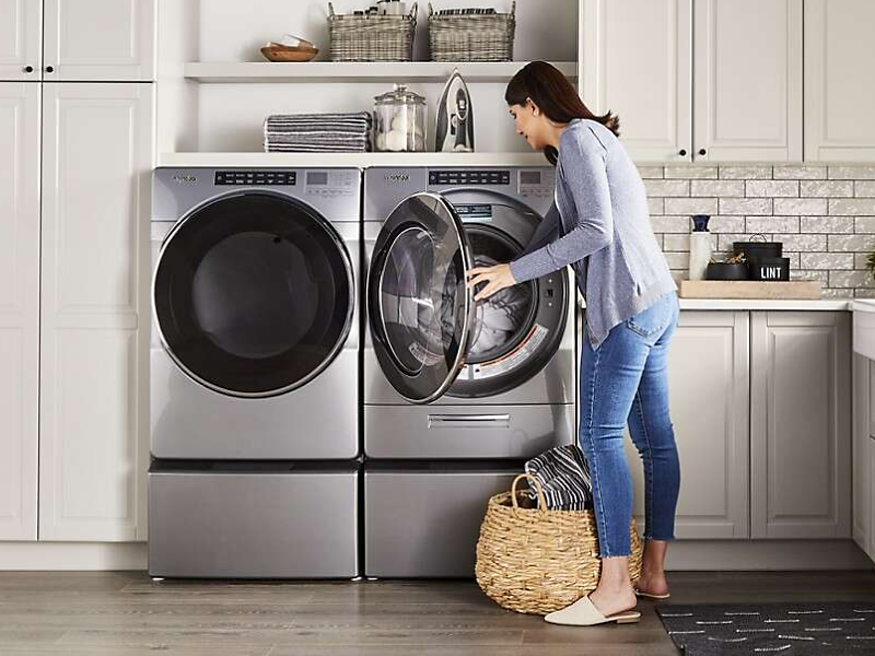 A woman putting laundry into a silver Whirlpool® front load dryer that sits next to a washer in a modern laundry room.