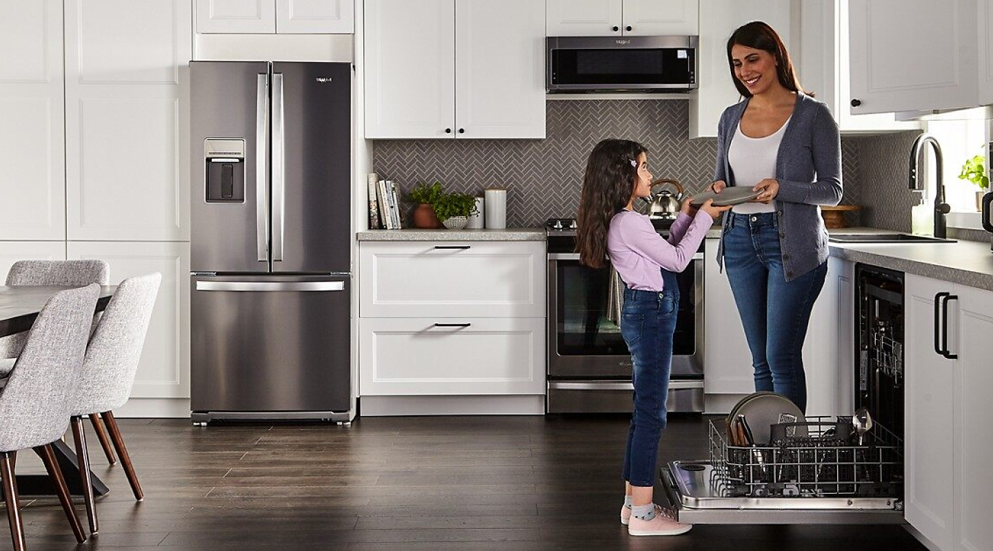 A woman handing a clean plate to a child next to a Whirlpool® dishwasher in a modern kitchen.A woman handing a clean plate to a child next to a Whirlpool® dishwasher in a modern kitchen.
