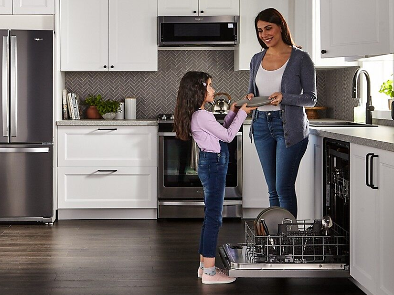 A woman handing a clean plate to a child next to a Whirlpool® dishwasher in a modern kitchen.