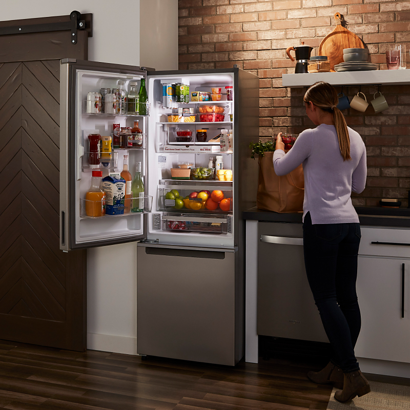A woman in a kitchen by an open refrigerator stocked with food