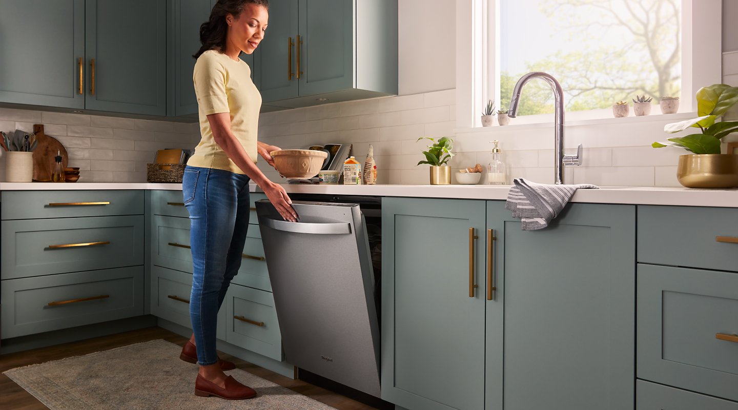 A person closing a stainless steel dishwasher surrounded by teal cabinetry.