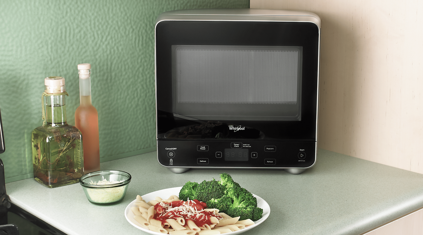A Whirlpool® Countertop Microwave on a kitchen countertop next to a plate of pasta and broccoli.