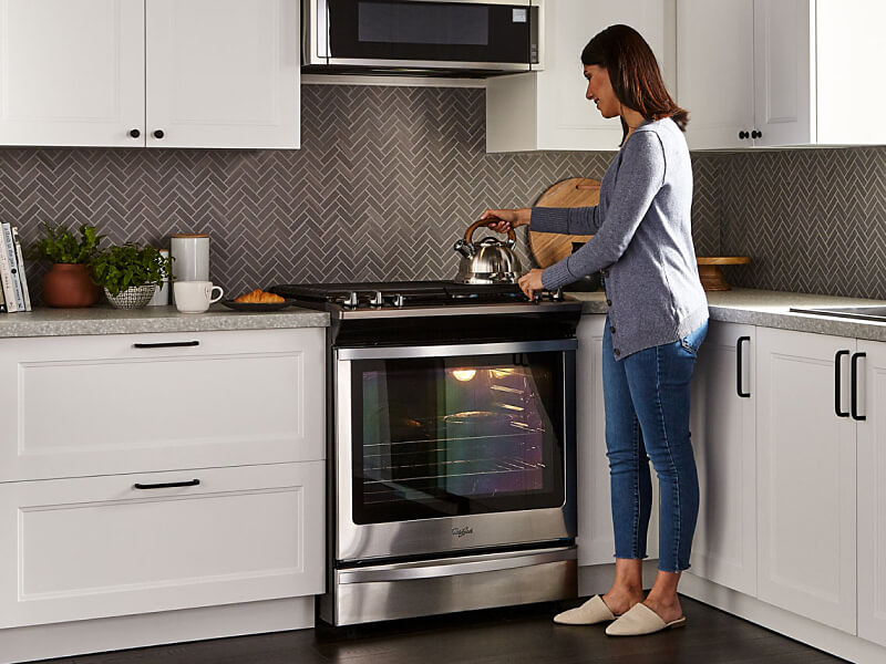 A person placing a tea kettle on the stovetop of a Whirlpool® gas oven in a modern kitchen.